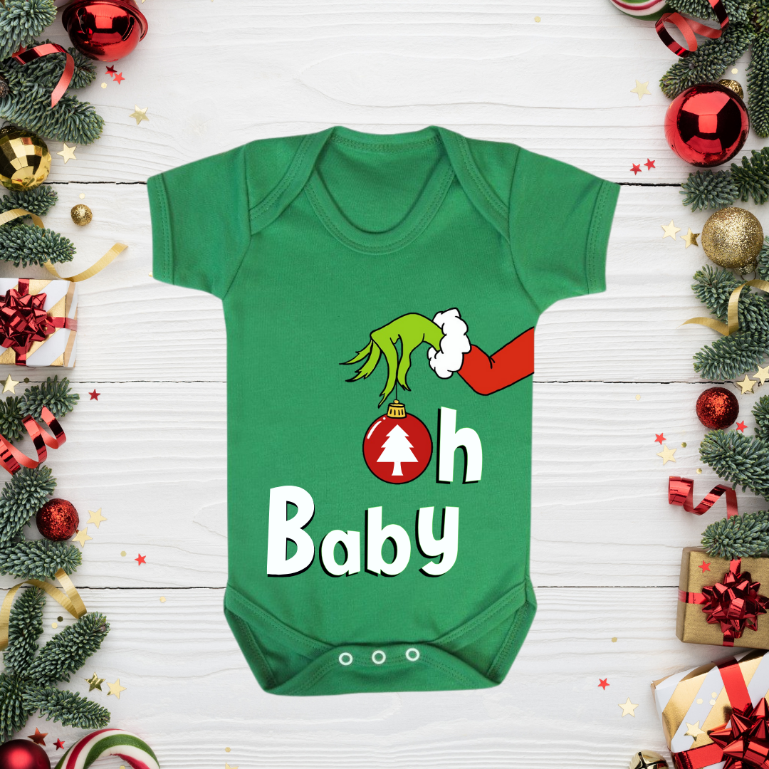 Green baby onesie with 'Grinch' design and 'Oh Baby' text, surrounded by Christmas decorations on a white wooden surface.
